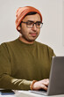 © TrueFrame Collective - Vertical medium shot of young Indian man wearing eyeglasses sitting at desk typing on laptop in office