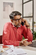 © TrueFrame Collective - Vertical medium shot of mature Indian manager sitting at desk in office having phone call with client