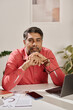 © TrueFrame Collective - Vertical medium portrait of mature South Asian man wearing coral shirt sitting at desk in office looking at camera