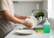 © Achmad - Cropped shot of a person in an apron washing a plate with a sponge under running water in a kitchen sink. Daily household chores and domestic hygiene concept.