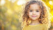 © UnPhouvith - A young girl with curly hair wearing a yellow sweater, standing outdoors with a blurred background of trees and sunlight.