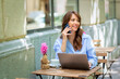 © sepy - Brunette haired woman sitting in an outdoor cafe and using smarpthone and computer laptop