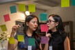 © Wasana - Two women are looking at a board with colorful sticky notes on it