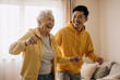 © Running opossum - Happy caucasian elderly woman and asian man dancing together in a cozy living room wearing yellow hoodies