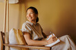 © BONNINSTUDIO/Stocksy - Smiling woman writing on notebook sitting on armchair at office