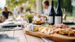 © dinatychynska - Outdoor wine and cheese tasting with artisan selection on wooden board. Elegant dining experience featuring gourmet bottles and fresh appetizers at restaurant terrace with people enjoying lunch.