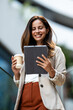 © Zoran Jesic - Smiling businesswoman using digital tablet and holding coffee outside office building