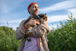© DimaBerlin - Happy man walking holding curious cat in tall summer grass, pet sniffing exploring surroundings. Pet owner with animal at outdoor adventure, feline curiosity stroll, bond with pet, nature exploration