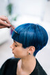 © Studio Marmellata - A professional hairdresser's hand carefully combs and styles the vibrant blue hair of a young woman. She has a trendy short haircut, receiving attention in a bright salon.