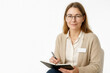 © Nargiz - Professional woman wearing glasses and a name badge, smiling warmly while holding a notebook and pen, seated against a white background, representing counseling, teaching, or professional consultation