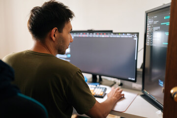 close-up rear view of programmer male working from home office using multiple monitors setup and typ