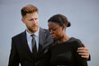 © Seventyfour - Caucasian young adult man comforting Black young adult woman wearing mourning veil outdoors during funeral ceremony, man placing arm around woman as she appears emotional