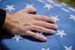 © Seventyfour - Caucasian middle aged man placing hand on folded American flag during funeral ceremony, close up of hand resting on flag with stars, symbolizing respect and mourning