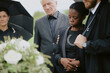 © Seventyfour - Black young adult woman standing with eyes closed being comforted by Caucasian middle aged man and woman during funeral ceremony outdoors near floral arrangement