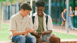 © Monkey Business - Two Male High School Or Secondary Students Looking At Social Media On Phone Sitting Outdoors