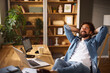 © Stockphotodirectors - A man sits comfortably in his home office, smiling and resting his hands behind his head. Natural light fills the cozy space, highlighting a desk with a laptop and a cup.
