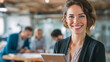 © Parol - Smiling woman in blazer holding tablet with colleagues working in the background in an office space