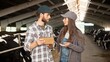 © VAKSMANV - Caucasian couple of young happy male and female farmers standing inside barn with cow flock, talking and using tablet device. Shepherds in stable with livestock having conversation typing on gadget