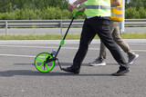 Two workers walking on road using measuring wheel for distance calculation.