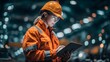 © CollyWang - Female worker in orange safety gear holding tablet in industrial setting.