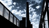 Silhouette of an industrial smokestack against a dramatic cloudy sky viewed from a modern urban setting