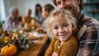 © junjie - Blond little girl smiling at camera while man in plaid shirt hugs her at Thanksgiving table with decorative pumpkin and autumn leaves, capturing warm and happy family gathering and festive gratitude