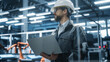 © Gorodenkoff - Factory Office: Portrait of Confident Caucasian Male Chief Engineer Wearing Hard Hat Working on Laptop Computer. Technician At Automated Robot Arm Assembly Line Manufacturing Electric Components.