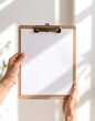 © Firla - Hands holding a wooden clipboard with a blank white paper mockup. Minimalist workspace scene with natural sunlight and shadows.