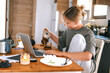 © shintartanya - Young woman working remotely at home kitchen table with laptop, coffee, and breakfast, representing freelance lifestyle, flexible schedule, and modern work-from-home concept.