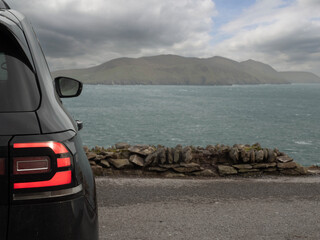 Wall Mural - Black car parked with view on amazing nature scene with ocean, mountain, and cloudy sky. Dingle peninsula, Ireland. Popular travel area with stunning Irish landscape. Nobody.