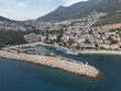 © MikeDrone - Kas harbor curves below steep hills in Antalya Province, Turkey, with a stone breakwater and small lighthouse, clear turquoise water and clustered white houses, calm midday light and gentle sea breeze