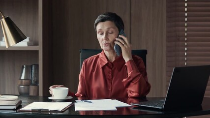 Wall Mural - Medium shot of mature Caucasian businesswoman talking on mobile phone drinking coffee while sitting in front of wireless laptop at office desk