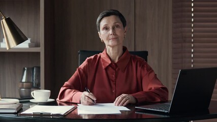 Wall Mural - Medium portrait of middle-aged Caucasian female entrepreneur doing report with financial documents sitting by laptop at office desk, smiling at camera