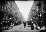 A historic black and white photo of a busy street with horse drawn carriages and people in vintage clothing