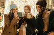 © Lomb - Three diverse friends drinking hot chocolate at a Christmas market at night. Happy multicultural women toasting with mugs of mulled wine, enjoying the festive holiday season.
