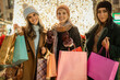 © Lomb - Three diverse friends with shopping bags looking at camera at a Christmas market. Happy multicultural women enjoying holiday gift shopping at night with festive lights.