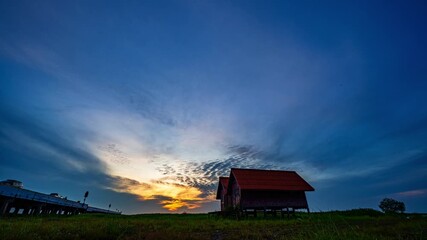Wall Mural - Time lapse Scenic view of two rustic lakeside houses with red roofs silhouetted against a dramatic sunset sky, with vibrant clouds spreading across the horizon and soft grass in the foreground.
