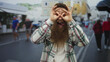 © Krakenimages.com - Young man with long beard makes binocular gesture with hands over eyes on sunlit busy street; curiosity.