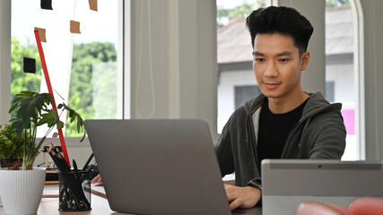 a focused asian creative freelancer man works on a laptop, developing graphic content from an office