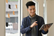© saltdium - A Smiling Asian man creating ideas on a digital tablet near a window, planning projects with adhesive notes
