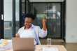 © MINAE - Young african american businesswoman raising her arms in celebration of achieving business goals using laptop, expressing joy and excitement in modern office workspace
