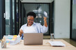 © MINAE - African american businesswoman raises her arms in excitement as she achieves a goal while working on her laptop, embodying success and achievement in a modern office environment