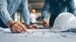 © Ivan - Construction workers are collaborating on detailed architectural plans at a building site. A hard hat is placed on the table as they mark key sections with pencils
