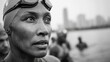 © Justlight - Focused medium shot on a participants concentrated face wearing a swim cap with the hazy outline of an urban waterfront and fellow swimmers in the background.