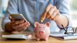 © NikProArt - Individual Saving Coins in a Piggy Bank While Managing Personal Finances Using a Smartphone and Calculator at a Desk