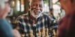 © Julian  - An older Black man laughs heartily while playing chess with a younger woman. They are seated at a wooden table in a warmly lit cafe. A third person, an older white woman