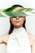 © VICHIZH - Portrait of a serene woman holding a large green leaf, embodying tranquility and nature in a minimalist white dress against a bright background.