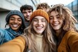 © smalltinykid - Group of five diverse young friends smiling and taking a selfie outdoors on a cloudy day, wearing winter clothes and looking happy and joyful