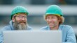 © BOJOShop - Two construction workers in green hard hats looking at a laptop screen and smiling