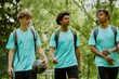 © Seventyfour - Three teenage boys, two Black and one Caucasian, walking outdoors with backpacks and soccer ball, engaging in conversation after sports practice in green park setting